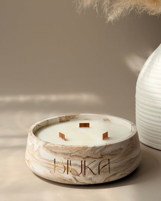 A round marble-look candle holder with three wicks and the brand name 'HUKA' printed on the front, styled on a beige surface with dried pampas grass and a white ribbed vase in the background.
