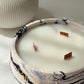 A marble-patterned candle bowl with white wax and three wooden wicks, featuring gold lettering that reads 'BIUKA', set against a neutral background with a ribbed white vase in the background.