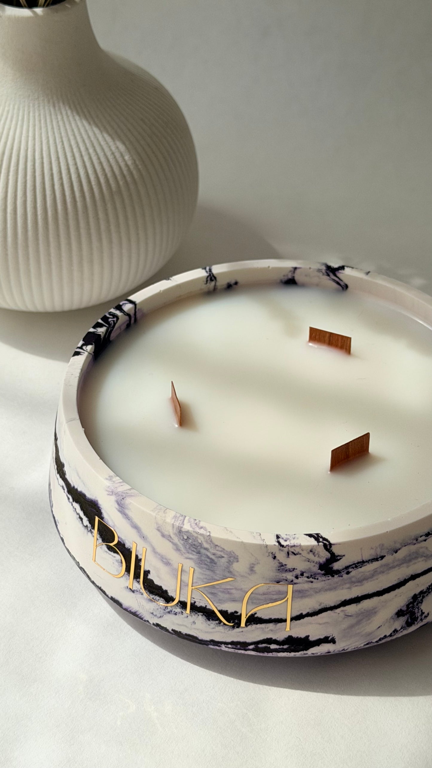 A marble-patterned candle bowl with white wax and three wooden wicks, featuring gold lettering that reads 'BIUKA', set against a neutral background with a ribbed white vase in the background.