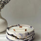 A round white candle with a WHITE and purple marble-patterned base and gold lettering, styled on a light gray surface next to a white vase with dried lavender sprigs.