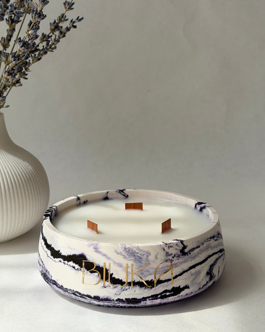 A round white candle with a WHITE and purple marble-patterned base and gold lettering, styled on a light gray surface next to a white vase with dried lavender sprigs.