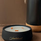 A black ceramic candle holder with gold script reading 'Santhalwood' on the front, filled with white wax and three orange wicks, styled on a brown surface next to a matte black vase with dried pampas grass.