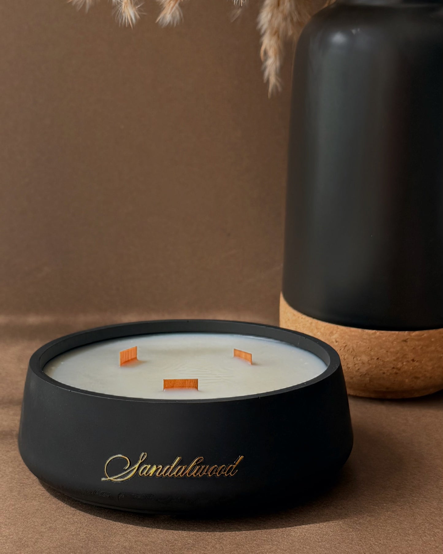 A black ceramic candle holder with gold script reading 'Santhalwood' on the front, filled with white wax and three orange wicks, styled on a brown surface next to a matte black vase with dried pampas grass.