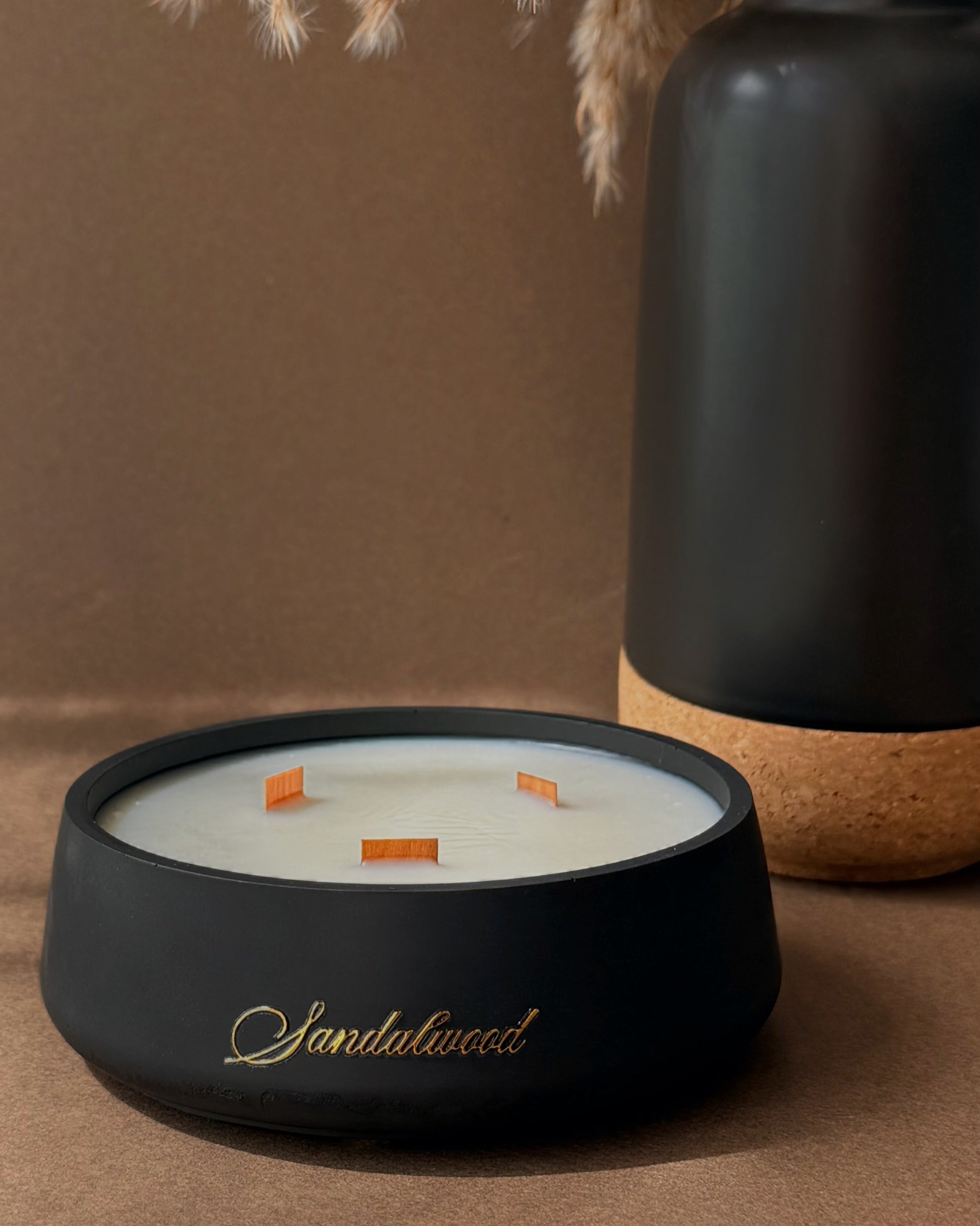 A black ceramic candle holder with gold script reading 'Santhalwood' on the front, filled with white wax and three orange wicks, styled on a brown surface next to a matte black vase with dried pampas grass.