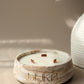 A round marble-look candle holder with three wicks and the brand name 'HUKA' printed on the front, styled on a beige surface with dried pampas grass and a white ribbed vase in the background.