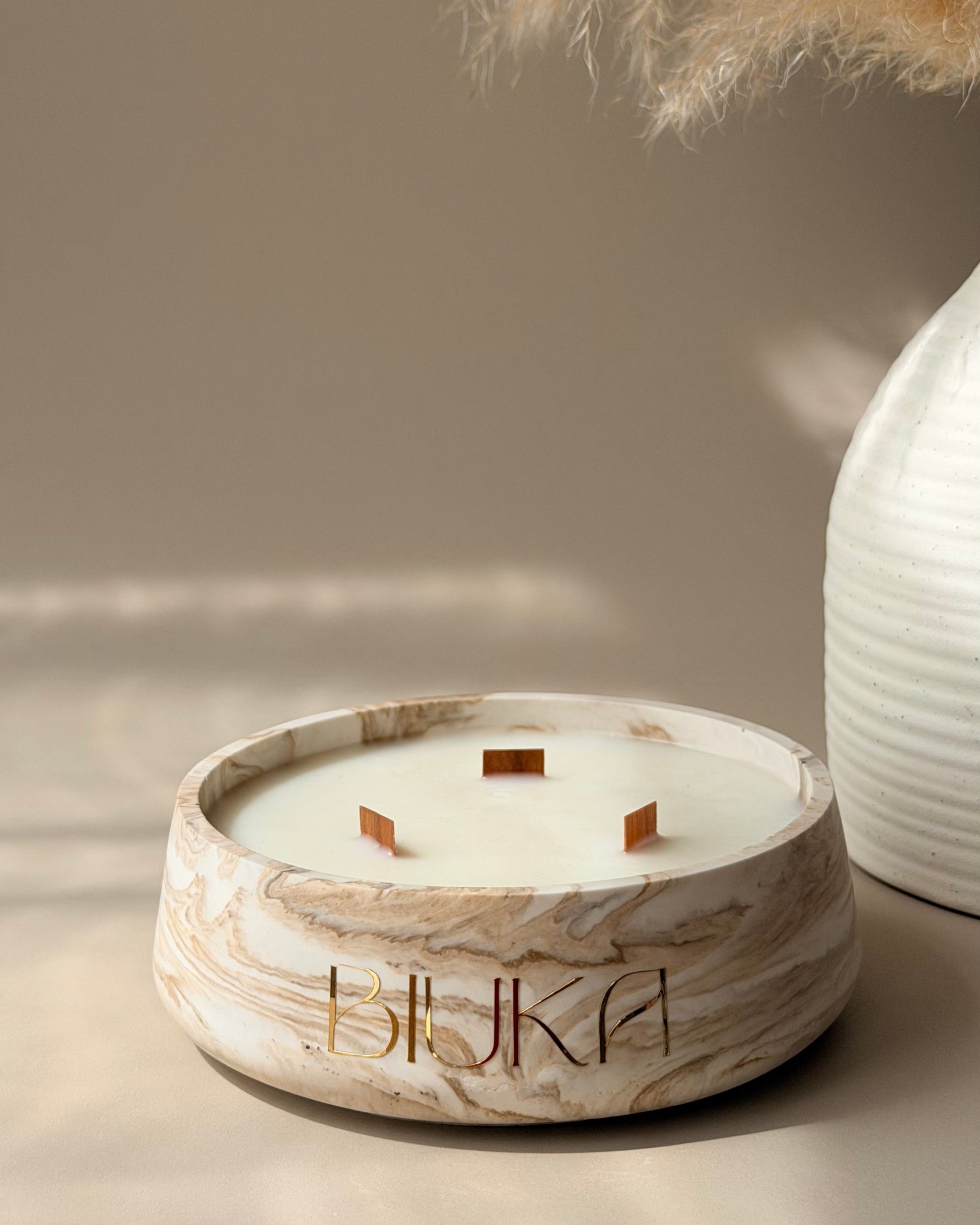 A round marble-look candle holder with three wicks and the brand name 'HUKA' printed on the front, styled on a beige surface with dried pampas grass and a white ribbed vase in the background.
