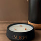 A matte black candle jar with the brand name 'BIUKA' in gold lettering, filled with white wax and featuring three small orange wicks. The candle is styled on a brown surface next to a black ceramic vase with dried pampas grass.