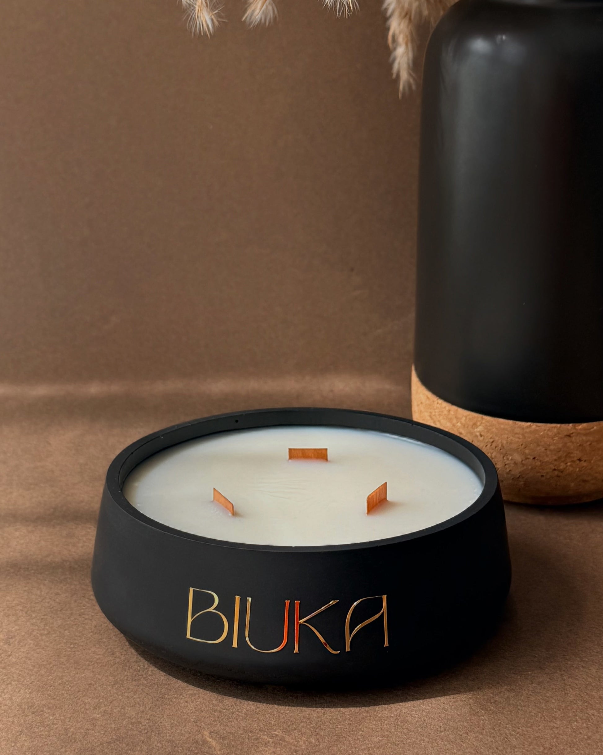 A matte black candle jar with the brand name 'BIUKA' in gold lettering, filled with white wax and featuring three small orange wicks. The candle is styled on a brown surface next to a black ceramic vase with dried pampas grass.