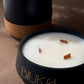 A close-up of a BIUKA candle in a matte black container with gold lettering, shown next to its open lid which reveals white wax and three wooden wicks. The candle is set on a textured brown surface with a dark background.