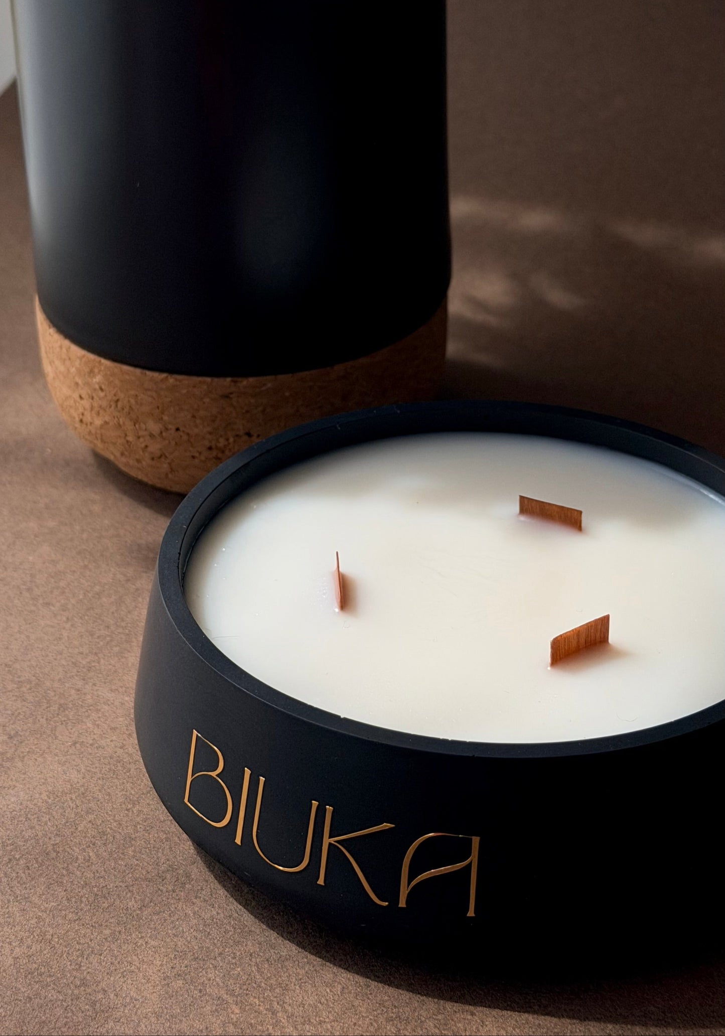 A close-up of a BIUKA candle in a matte black container with gold lettering, shown next to its open lid which reveals white wax and three wooden wicks. The candle is set on a textured brown surface with a dark background.