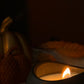 A close-up of a lit candle in a matte black holder with gold lettering, set against a dark, warm-toned background with soft decorative elements.