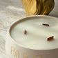 A close-up of a round white candle with three wicks and the brand name 'BIUKA' in gold lettering on the front. The candle is set on a light marble surface with a blurred gold decorative object in the background.