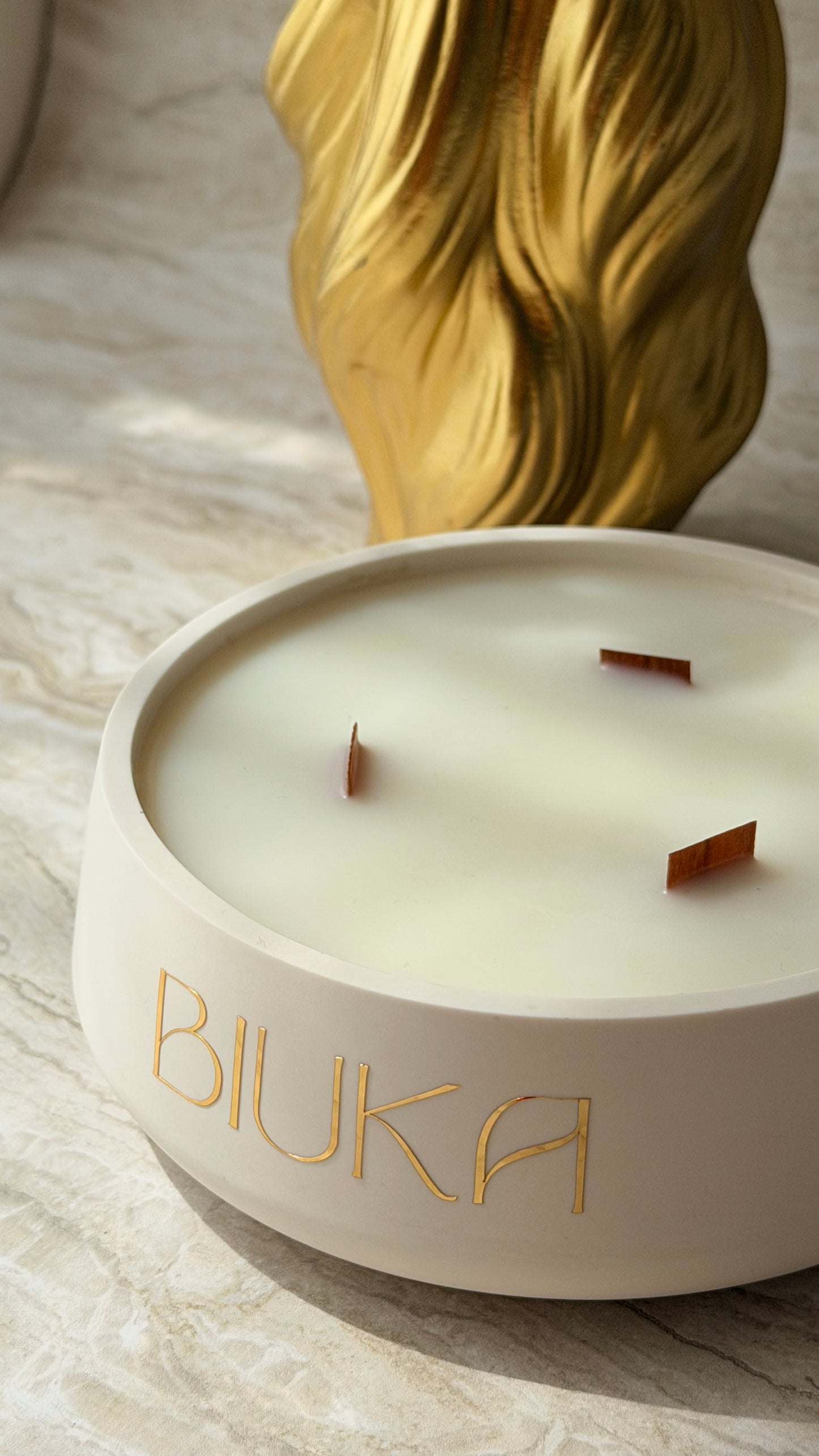 A close-up of a round white candle with three wicks and the brand name 'BIUKA' in gold lettering on the front. The candle is set on a light marble surface with a blurred gold decorative object in the background.