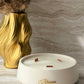 A white ceramic scented candle in a shallow round dish with three wicks, featuring the brand name 'Blossom' in gold script. The candle is styled on a light marble surface next to a gold vase with dried grass.