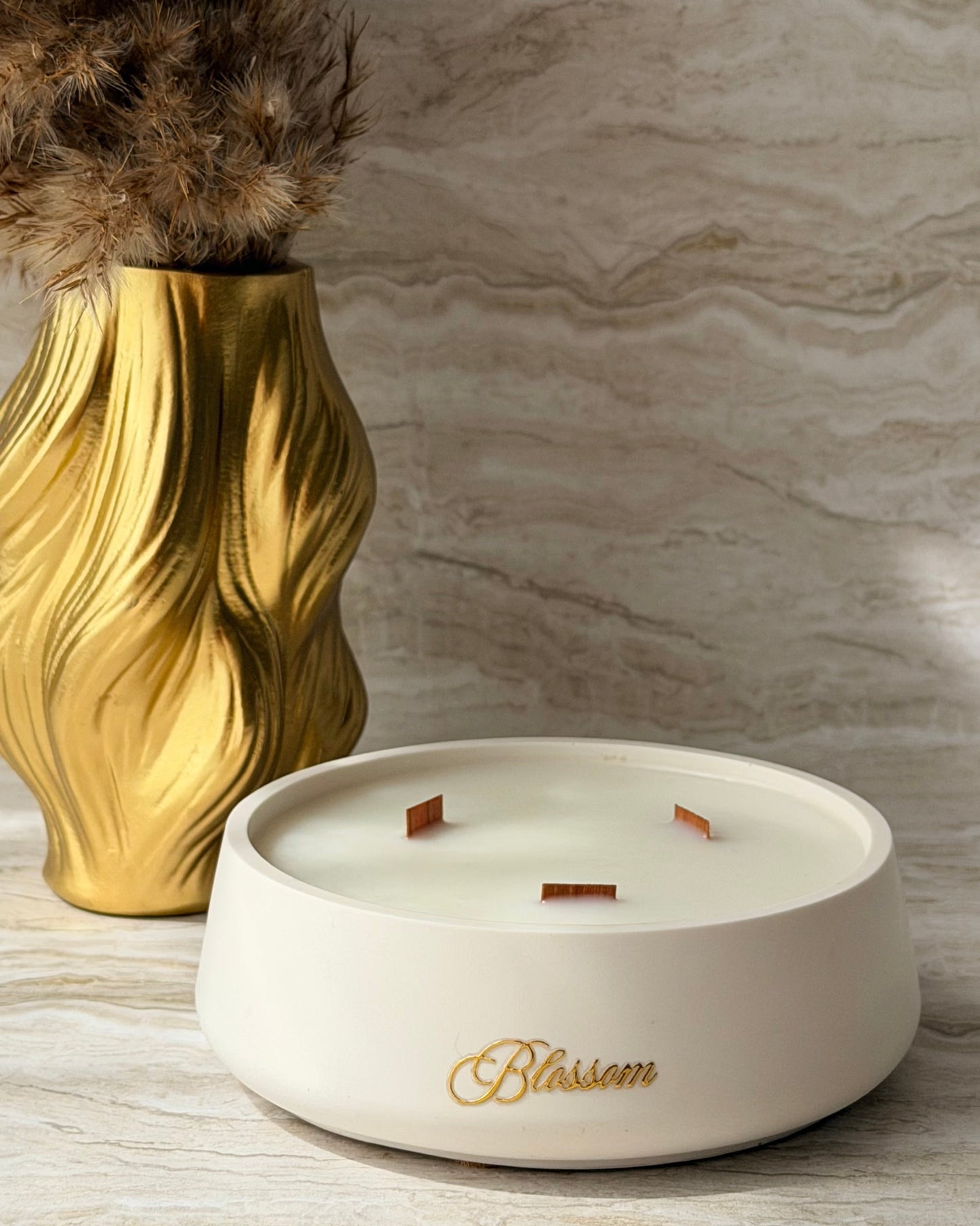 A white ceramic scented candle in a shallow round dish with three wicks, featuring the brand name 'Blossom' in gold script. The candle is styled on a light marble surface next to a gold vase with dried grass.