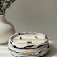 A round marbled candle in a white ceramic holder with black and purple veining, featuring three wicks and a gold script logo, styled on a light surface next to a white ribbed vase with dried lavender.