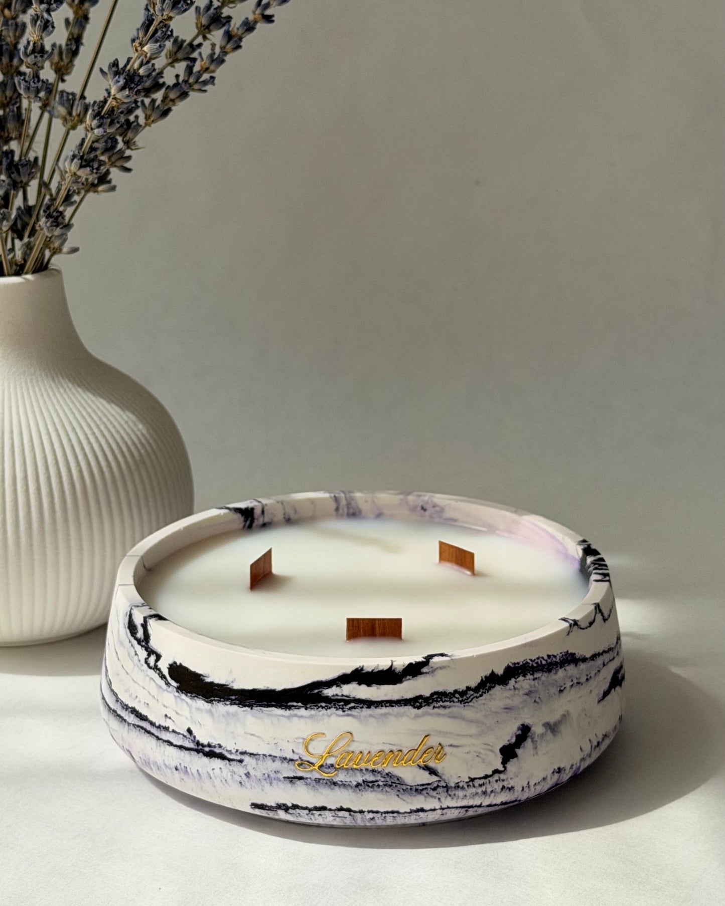 A round marbled candle in a white ceramic holder with black and purple veining, featuring three wicks and a gold script logo, styled on a light surface next to a white ribbed vase with dried lavender.