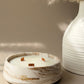 A round white and beige marble-patterned candle holder with three wicks and the word 'Cinnamon' in gold script, styled next to a white ribbed vase with dried pampas grass against a warm beige background.