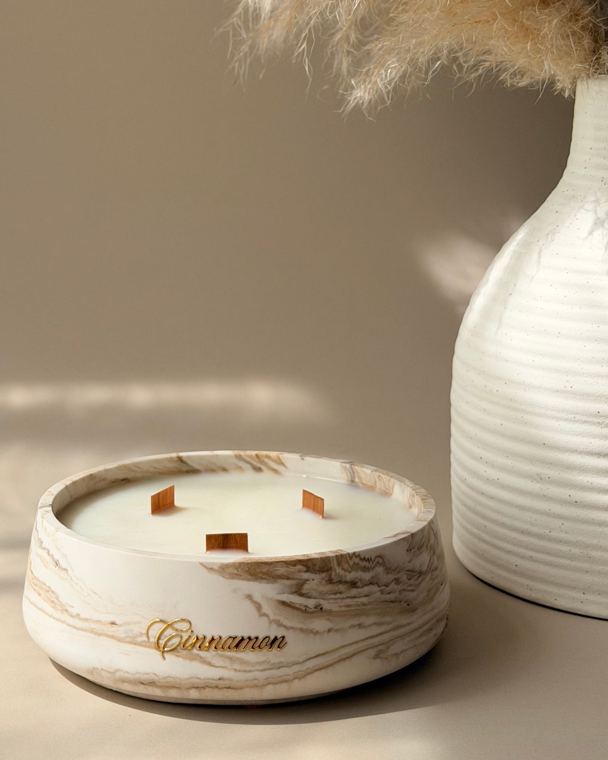 A round white and beige marble-patterned candle holder with three wicks and the word 'Cinnamon' in gold script, styled next to a white ribbed vase with dried pampas grass against a warm beige background.