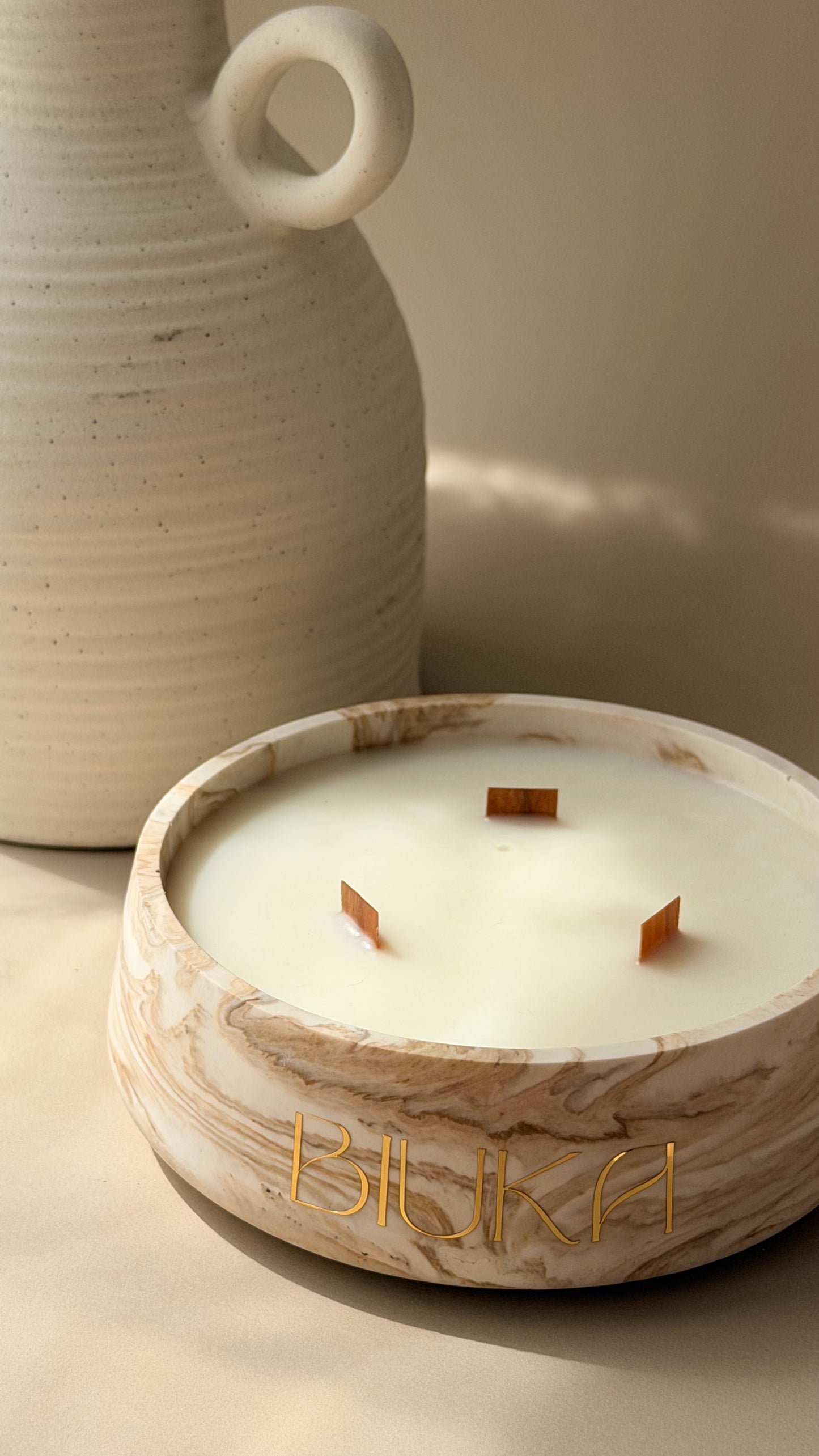 A close-up of a marble-look candle bowl with three wicks and the brand name 'BIUKA' in gold lettering, set against a warm beige background with a ceramic vase partially visible.