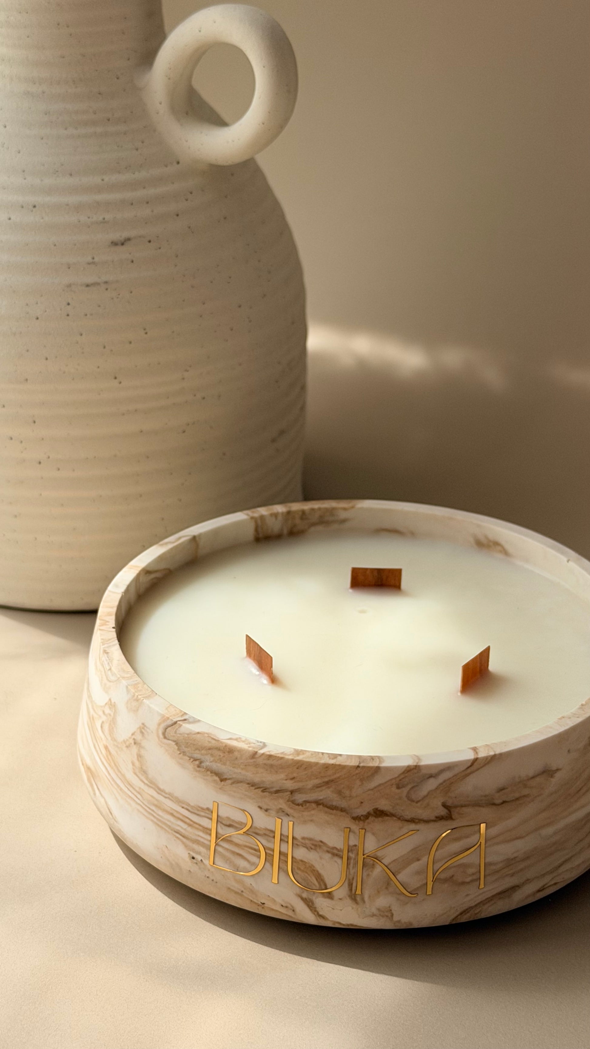A close-up of a marble-look candle bowl with three wicks and the brand name 'BIUKA' in gold lettering, set against a warm beige background with a ceramic vase partially visible.