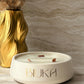 A cream-colored ceramic candle holder with the brand name 'BIUKA' printed in gold on the front, styled on a marble surface next to a gold vase with dried grass.