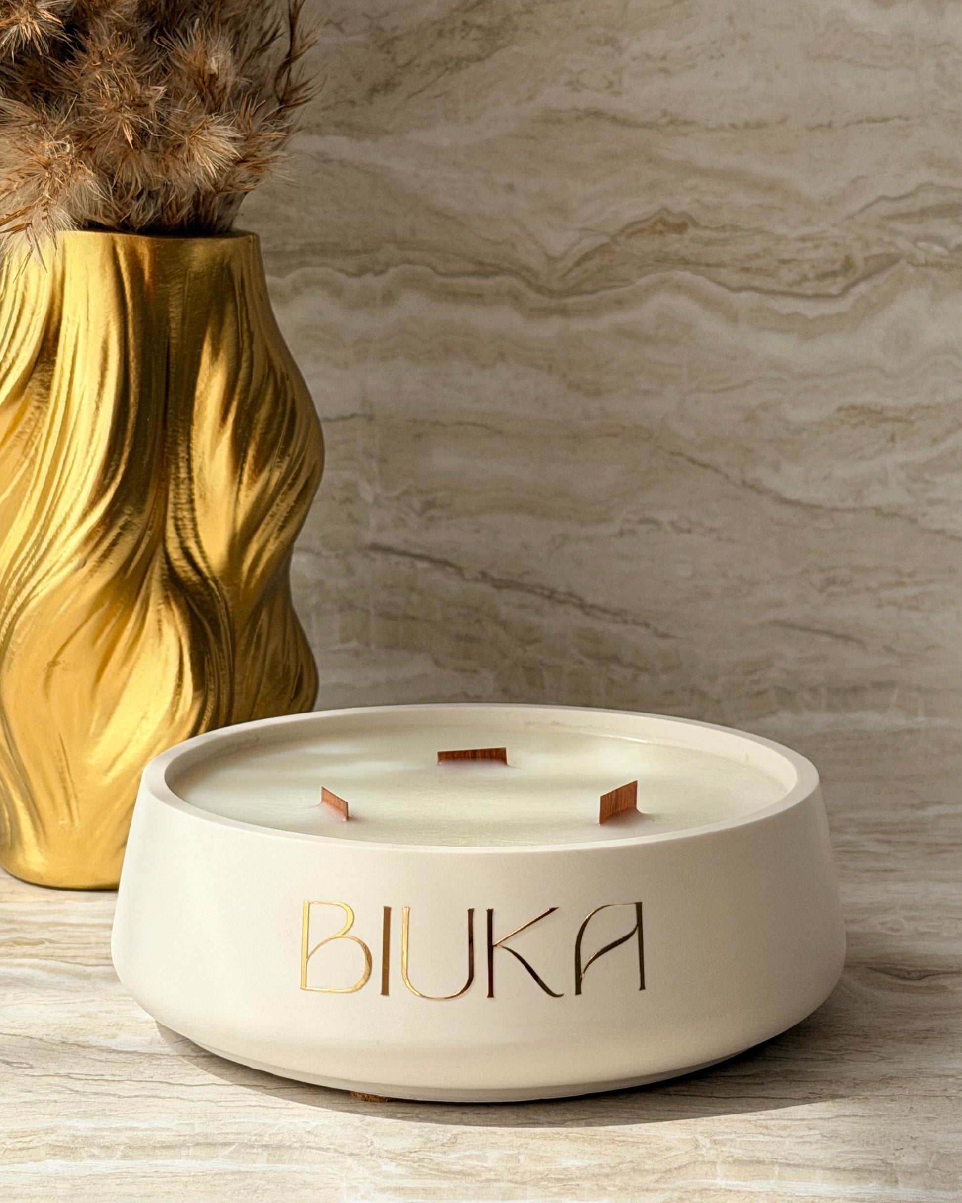 A cream-colored ceramic candle holder with the brand name 'BIUKA' printed in gold on the front, styled on a marble surface next to a gold vase with dried grass.